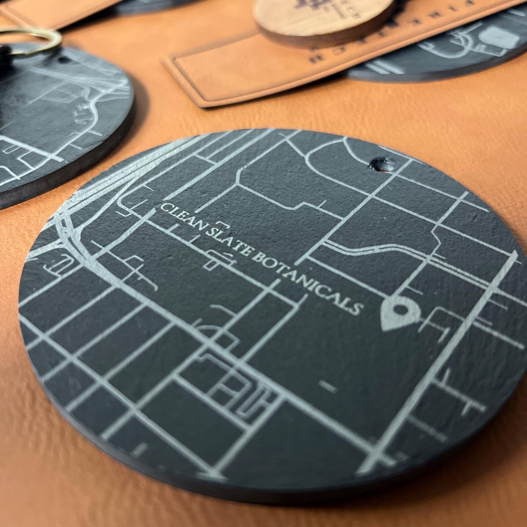 Round dark green coaster with map design and 'Clean Slate Botanicals' text on a brown leather surface.


