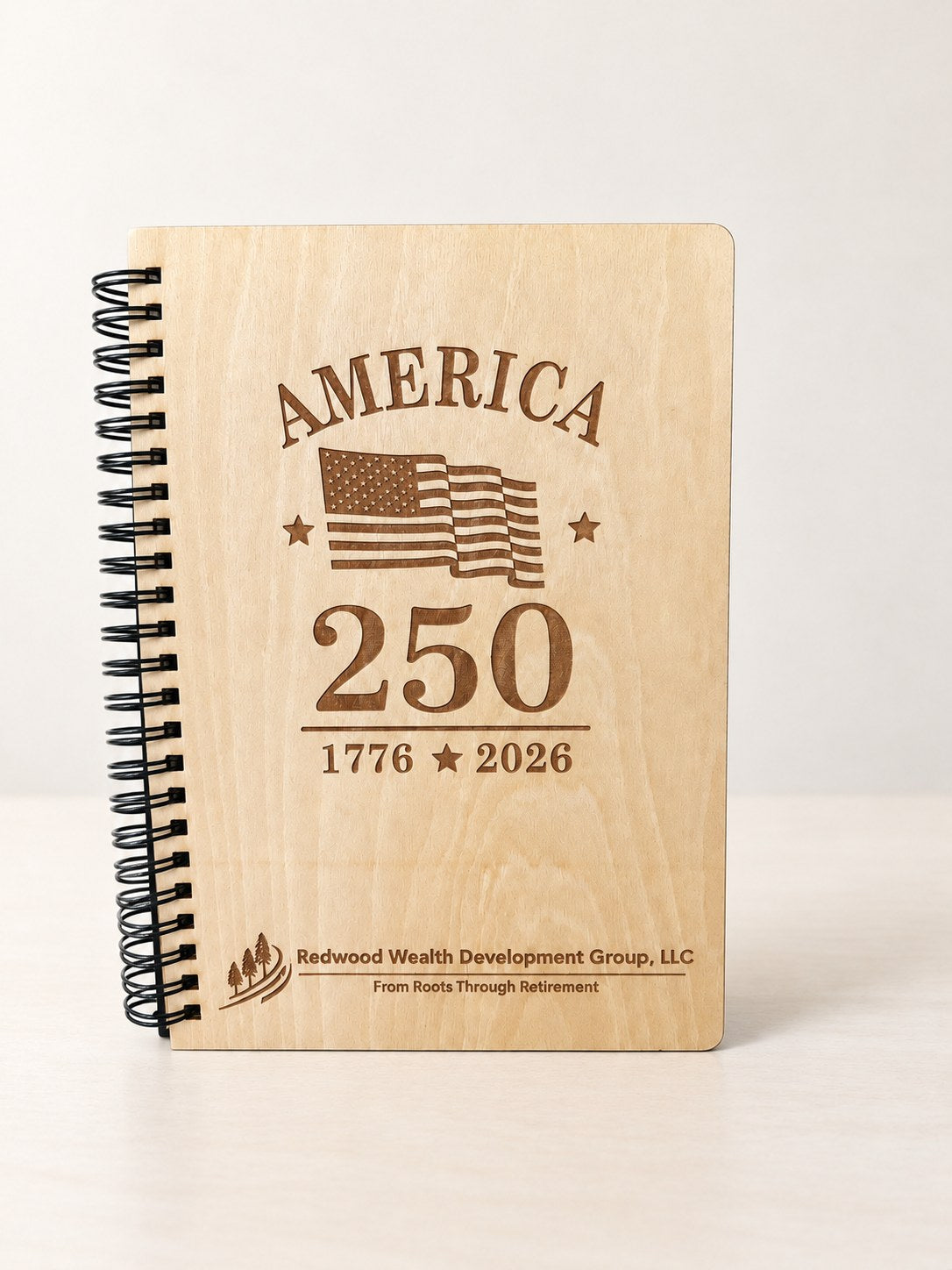 Wood spiral notebook with a black spiral standing on a table, laser engraved with America 250, a flag and a company logo